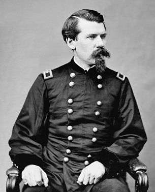 A white man with a Van Dyke beard and mustache sitting in a chair, wearing a baggy double-breasted military jacket.