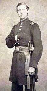 A white man in a long military jacket, with a wide sash and belt at the waist and a sword hanging at his side. He is standing with his right hand inside his jacket.