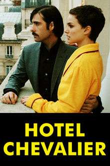 A young white couple stand on a hotel balcony looking out to the left of the frame. Only their upper bodies are shown. The man, who has shoulder-length hair and a trim moustache and who is wearing a grey suit, has his left arm around the short-haired woman, who is clad in a yellow hotel bathrobe and has her right hand on his shoulder.