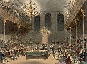 View of a large room, showing the Speaker of the House sitting at the end. Down each side of the room, MPs are sitting – one MP is standing on the right, giving a speech. Balconies are on either side, with spectators visible.