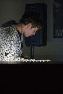 A man in his twenties with short messy brown hair concentrates on the decks in front of him with headphones tucked above his ear whilst wearing a white and black patterned hoody.