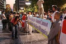 A crowd of protestors along a street displays a banner reading "Human Rights Abuse Cannot Co-exist with Beijing Olympics". Near the centre of the image, a photographer holds a camera level with the banner while looking through the viewfinder.