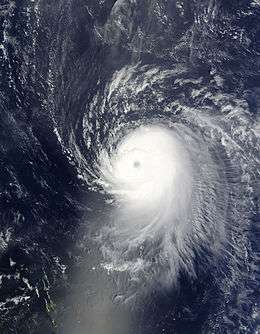 Satellite image of a slightly elongated tropical cyclone over blue waters; an eye, visible as a void at the center of the mass of white clouds, is visible. Small green islands dot the lower-left corner of the image.