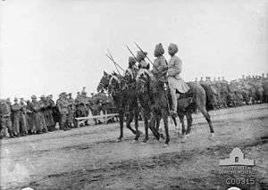 In a black and white photograph, four men with swords raised at 45 degrees wearing military uniforms and turbans ride on dark-coloured horses facing left. Behind the men, a large crowd of soldiers look on.