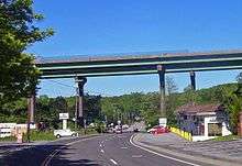 A pair of bridges pass from left to right on tall supports that tower over the other structures in the area, such as telephone poles and single-story buildings. The two-lane NY 22 passes underneath the bridge and proceeds into the background.