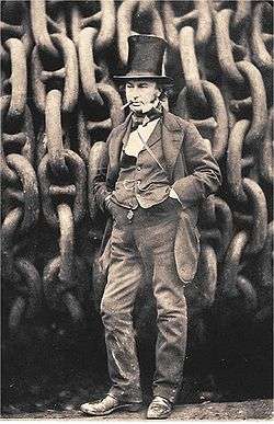 A 19th century man wearing a jacket, trousers and waistcoat, with his hands in his pockets and a cigar in mouth, wearing a tall stovepipe top hat, standing in front of giant iron chains on a drum.