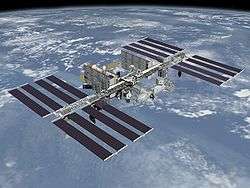 A planform view of the International Space Station is backdropped by the limb of the Earth. In view are the station's four large, blue-colored solar array wings, two on either side of the station, mounted to a central truss structure. Further along the truss are six large, white radiators, three next to each pair of arrays. In between the solar arrays and radiators is a cluster of pressurised modules arranged in an elongated T shape, also attached to the truss.
