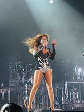 The image of a woman who is singing. She wears a black leotard and holds a microphone with her right hand while she moves quickly her left hand. Many musical instruments are visible behind her.