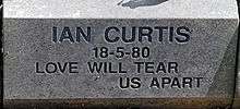 A grayish stone block with "Ian Curtis 18-5-1980 Love Will Tear Us Apart" carved into it in a sans-serif typeface