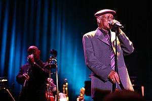 A man wearing a cap on his head, eyeglasses, and a suit, holding on to a microphone stand with both hands. To the left of him is a man also holding on to a microphone stand. In between them are various musical instruments.