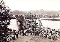 An old photograph showing a crowd of people in the foreground with a steel bridge spanning a river in the background