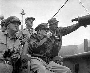 MacArthur is seated, wearing his field marshal's hat and a bomber jacket, and holding a pair of binoculars. Four other men also carrying binoculars stand behind him.