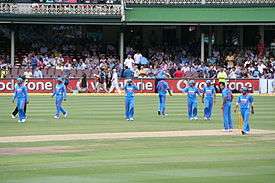 The view of a cricket field. Players wearing blue outfits can be seen.