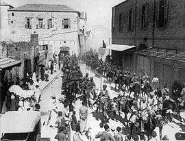In a black and white photograph, a large group of turbaned men on horseback ride through a dusty, sunlit street and into the distance, obscured by dust. A crown of civilians watch the men pass. A large, brick-built, two-storey building is on the left, and a similar structure is on the right.