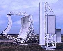 Colour photograph of two large white-painted items of equipment (the left being a parabolic 'dish' with straight sides; the right a tall, narrow structure) on grass, with cloudy sky above.