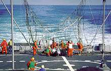The rear deck of ship, with a large partially erect net visible near the center of the image. Many men in orange suits are working to free a white drone entangled in the net.