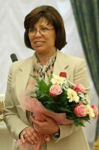 A woman with brown hair, wearing eyeglasses and a tan suit, holding a bouquet of flowers.