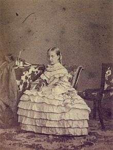 A photograph of a young, light-haired Isabel wearing an elaborate dress with a layered, hooped skirt and seated in front of a table that holds several books