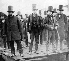 A group of ten men in nineteenth century dark suits, wearing top hats, observing something behind the camera