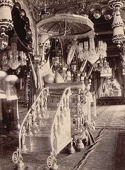 A black and white picture of an ornamented throne, constructed out of some metal. There are intricate carvings of horses on its base and of women on the steps leading up to it. An umbrella rises above the throne and many chandeliers surround it.
