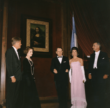 Three men and two women stand near the Mona Lisa. All are dressed formally, one woman in a spectacular pink gown.