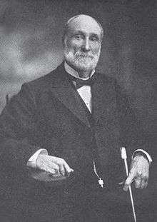 A man with receding gray hair and a gray beard and mustache. He is wearing a black coat, white shirt, and black bow tie