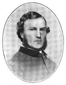 Head of a white man with wavy hair and a beard but no mustache, wearing a stiff-collard jacket.