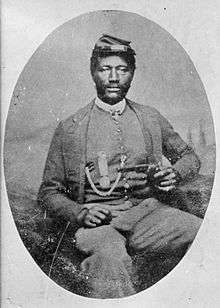 A black man sitting in a chair, wearing a forage cap and a suit coat over a vest and holding a revolver in his left hand. On the vest is a watch fob chain and a circular medal hanging from a ribbon.