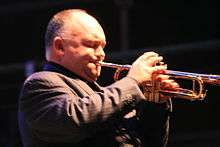 A 45-year-old man is shown in right profile, he is playing a trumpet with his right hand manipulating the valves and his left holding the instrument to his pursed lips. He has sparse thin head hair, his eyes are closed and he wears a brown pin-strip suit jacket. The front of the trumpet is out of shot.