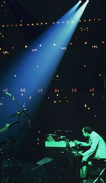 To one side of a stage, a man plays keyboards while bathed in green light