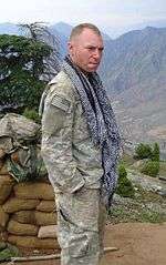 A white man with close-cropped blond hair standing with his hands in his pockets, wearing a camouflage uniform and a long blue and white scarf hanging untied around his neck. Behind him are a wall of sandbags, a tree and, in the distance, mountains.