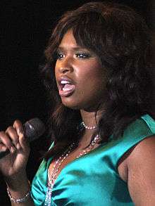 A black women with dark brown hair talking into a microphone. She is wearing a green satin dress with a with necklace.