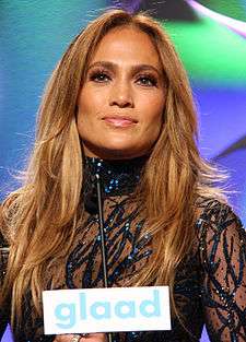 A portrait of Jennifer Lopez posing onstage in a black see-through outfit.