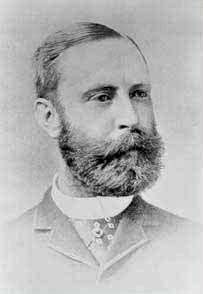 A black-and-white photographic portrait of a man with a thick beard and mustache wearing a high collar and jacket. It is faded around the edges.