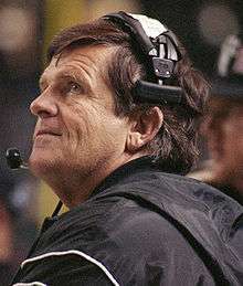 Head shot of a coach with brown hair wearing a headset and black jacket.