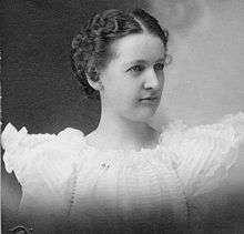 Head and shoulders of a woman, about age 20. Her dark hair is tightly coiffed and parted in the middle near her forehead. The ruffles of her white dress extend like stubby wings from her shoulders.
