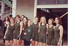 Nine smiling young women, eight in green one-piece tight lycra outfits and the shortest in jumper and trousers, stand in a line in front of two crossed oars