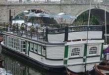 A barge moored near a large stone bridge, stern closest to the viewer, with a black hull and white cabin timbers with green trimmings. On the cabin roof, there are tables, chairs and parasols.