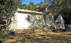Photograph of the Joaquin Miller House. A small house on a gradual incline, elevated by brick foundation, surrounded by trees.