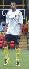 Man wearing white t-shirt, black shorts and yellow socks, standing on a pitch
