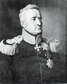 Black and white print shows a stern-looking man with short hair. He wears a dark military coat with a light-colored collar and epaulettes. An Iron Cross can be seen pinned to his coat.