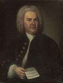 portrait of Bach towards the end of his life, posing in a dark outfit with a wig, holding a single sheet of music in his right hand, facing the viewer with a serious expression