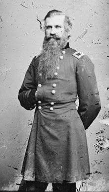 A white man with a full beard extending down to the center of his chest, wearing a long military jacket. He is standing with one hand behind his back and the other inside his jacket.
