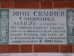 A tablet formed of six standard sized tiles, bordered by green flowers in the style of the Arts and Crafts movement. The tablet reads "John Cranmer Cambridge aged 23, a clerk in the London County Council who was drowned near Ostend whilst saving the life of a stranger and foreigner, August 8, 1901".