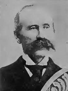 Head of a white man with a large mustache and receding hairline wearing a dark suit, white shirt, and dark tie. Over the bottom right of the portrait is a decorative design.