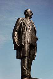 Bronze statue of Diefenbaker, taken from one side. He is depicted wearing an overcoat over a suit. He carries the Bill of Rights under his arm.
