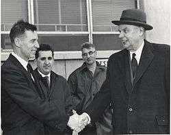 Diefenbaker, wearing a coat over his suit, shakes hands with a smiling man. Two other men, looking less impressed, are in the background.