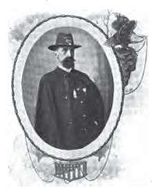 Head and torso of a white man with bushy beard wearing a wide-brimmed hat and a dark jacket. The right sleeve of the jacket is hanging empty.