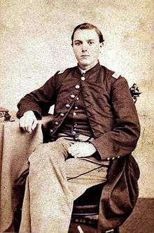 Young white man sitting on a chair with his legs crossed and his right arm resting on a table beside him. He is wearing light colored pants and a military jacket buttoned at the top only with a vest underneath.