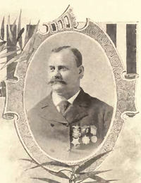 Head and shoulders of a white man with a mustache and receding hairline, wearing a dark suit and tie with three medals hanging from ribbons on the left breast. The portrait is surrounded by a decorative oval frame.